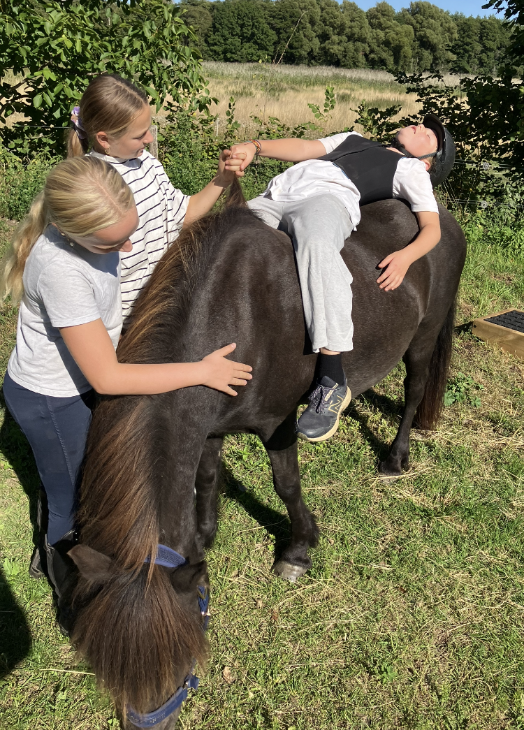 tre unge mennesker, hvor af en ligger på en hest, og holder en af de andre i hånden