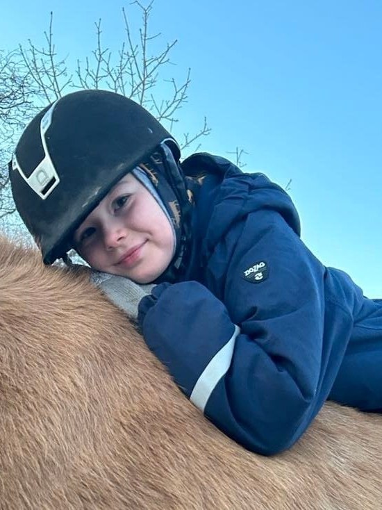 Barn med ridehjelm ligger smilende hen over ryggen på en islandshest under en klar blå himmel