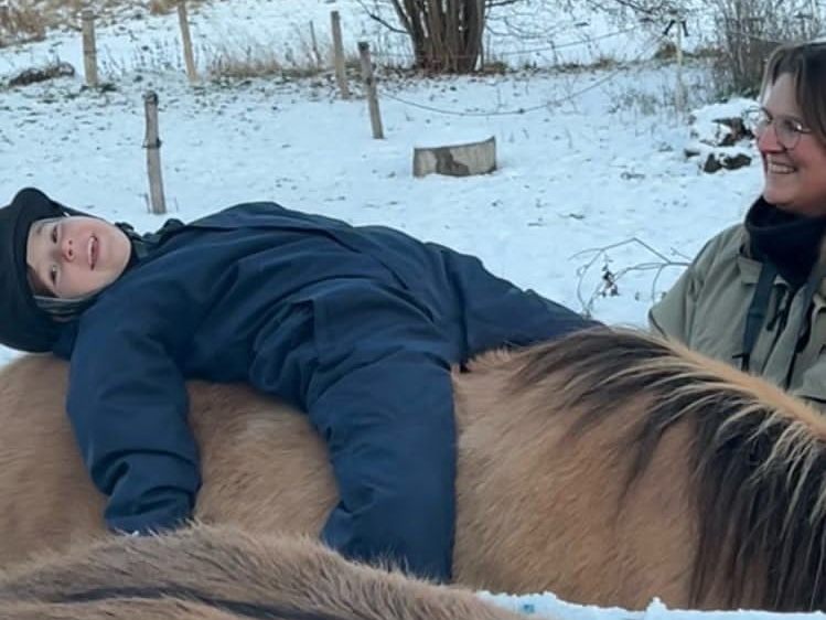 Barn ligger afslappet på tværs af en islandshests ryg, mens Lotte står smilende ved siden af