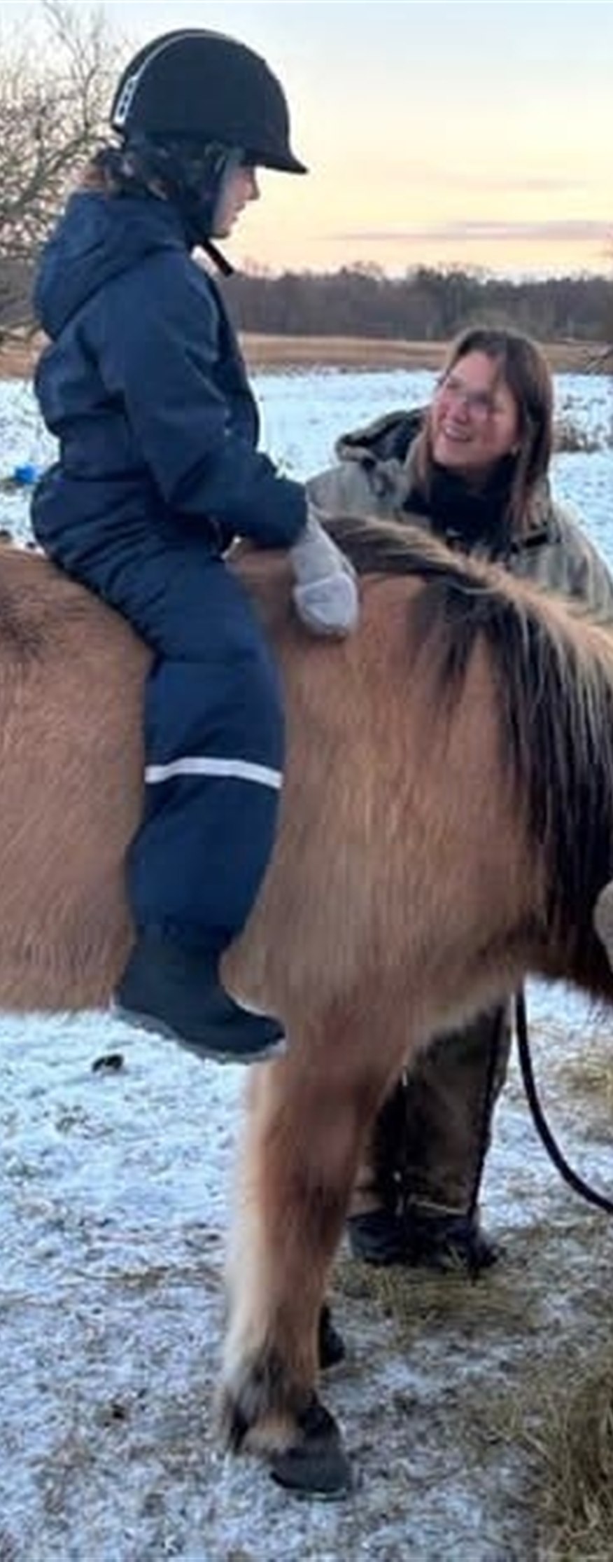 Barn med ridehjelm sidder på en islandshest, mens Lotte står smilende ved siden af i sneen