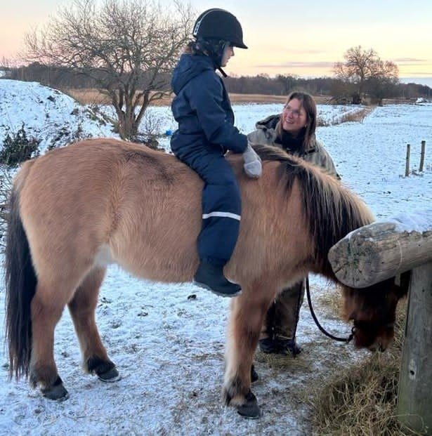 Barn med ridehjelm sidder på en islandshest, mens Lotte står ved siden af i sneen